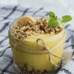 A glass of yellow pudding with nuts and banana.