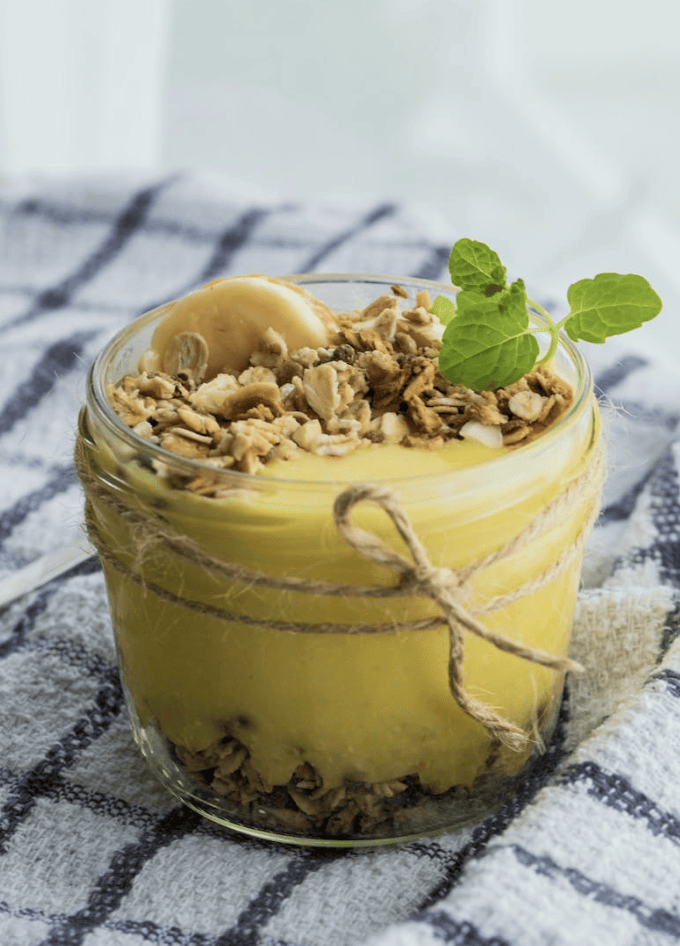 A glass of yellow pudding with nuts and banana.